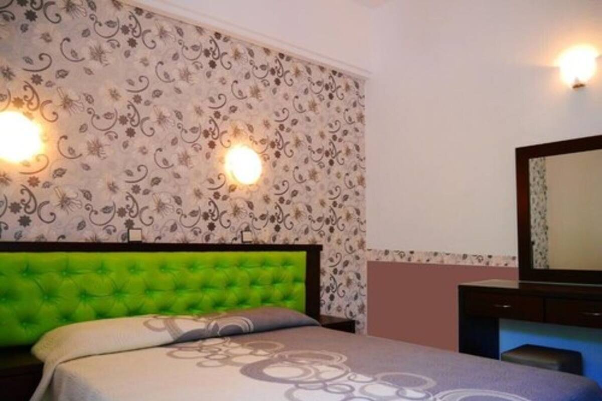 A hotel room with a green upholstered headboard, a patterned bedspread, and a wall-mounted mirror.