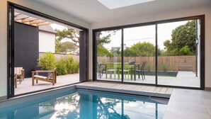 A modern indoor swimming pool with a skylight, large sliding glass doors, and a patio area with outdoor furniture. - Villa Odet O Bord de l'O Piscina coberta 29°C em Sainte Marine, praia 100m (Combrit)