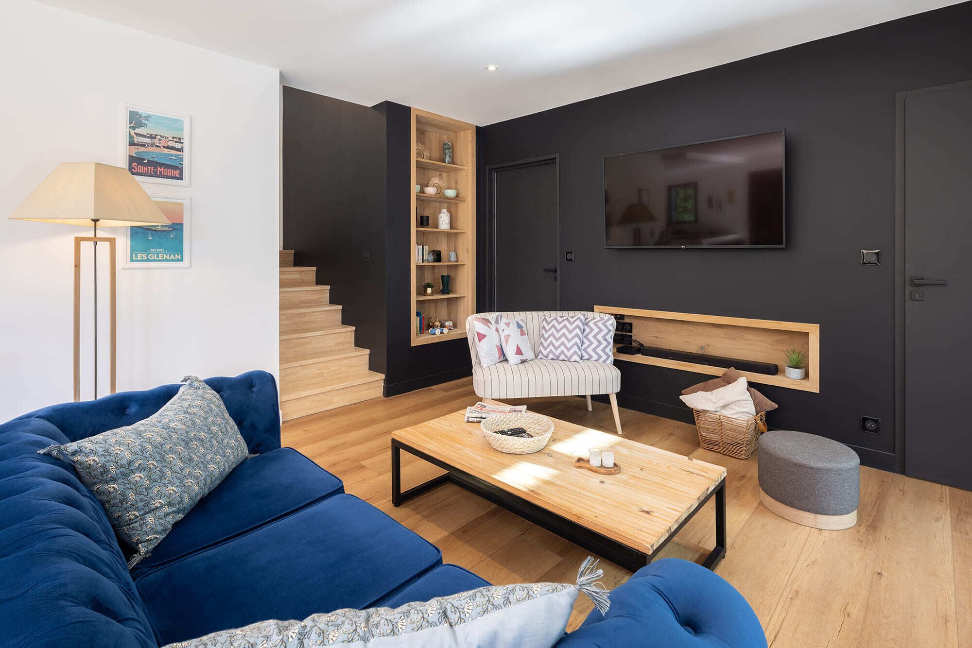 A modern living room with a blue sofa, a wooden coffee table, a bookshelf, and a TV mounted on the wall.