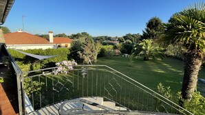 Un balcon donnant sur un jardin bien entretenu, un palmier et une maison avec un toit rouge en arrière-plan.