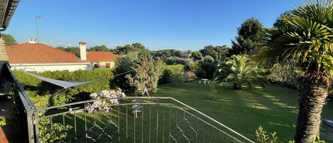 En balkon med udsigt over en velholdt have, et palmetræ og et hus med rødt tag i baggrunden.