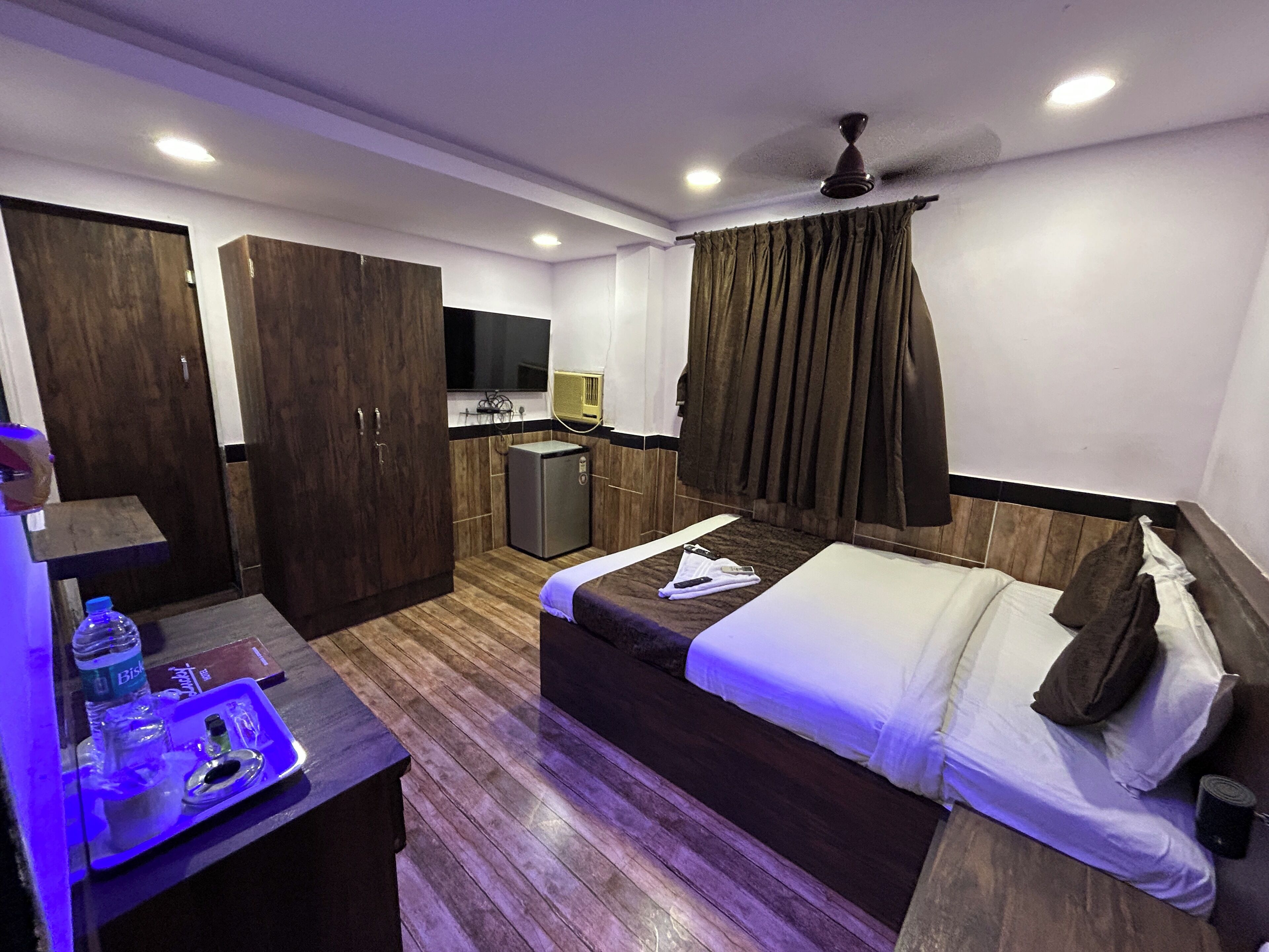 Habitación de hotel con cama, escritorio, una botella y vasos, kitchenette pequeña y ventilador de techo.