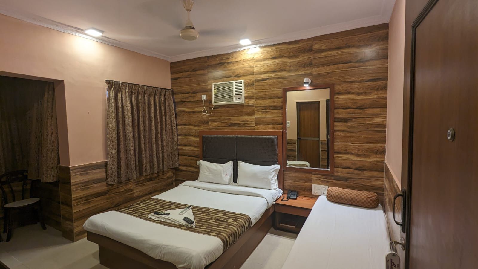 Un dormitorio con panel de madera en la pared, cama con ropa de cama blanca, mesita de noche con lámpara y espejo sobre la cama.
