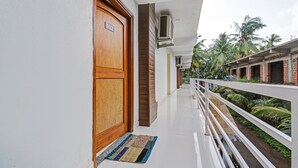 A corridor with a wooden door labeled "102", a white railing, and a view of a garden with palm trees. - Itsy Hotels Morjim Pride (Morjim)