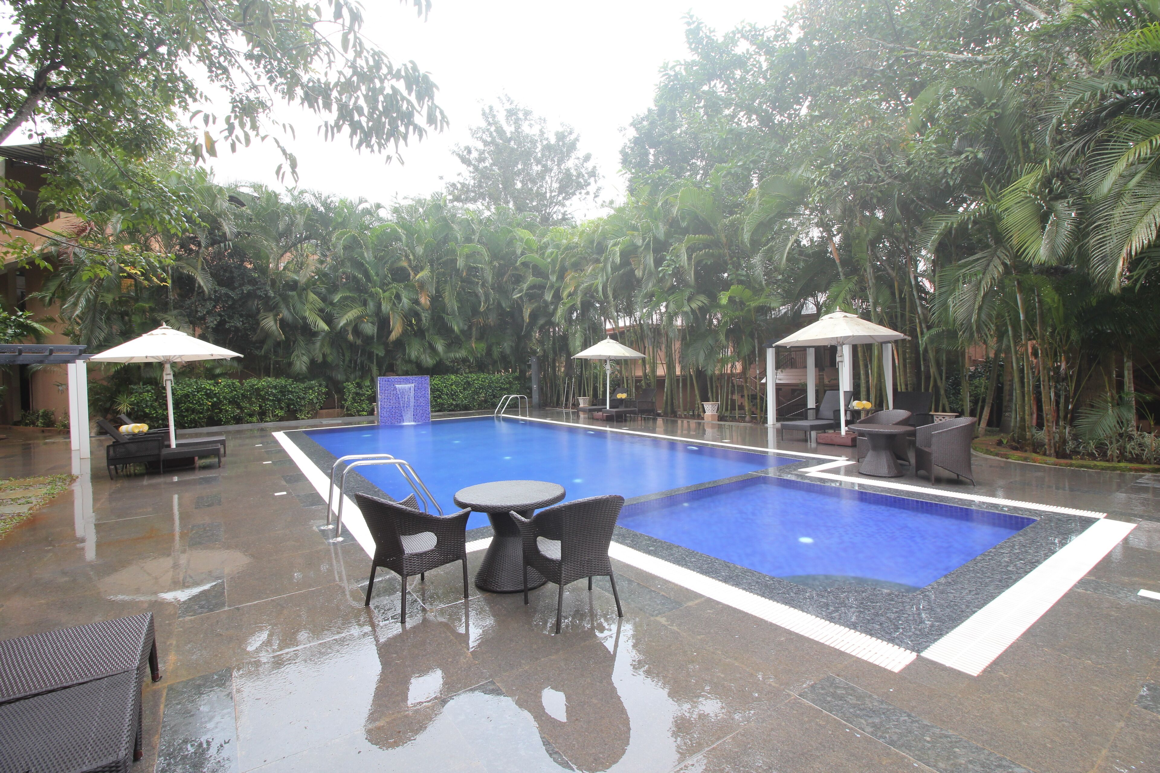 A swimming pool with lounge chairs and umbrellas, surrounded by palm trees and a tiled patio area.