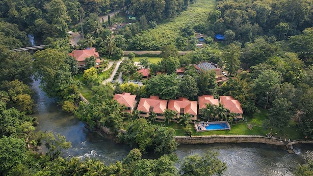 An aerial view of a resort with multiple red-roofed buildings, a swimming pool, and a river running through the property. - Regenta Jungle Resort Kabini Springs (Heggadadevankote)