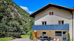 A two-story building with a balcony, a paved driveway, and a mountain backdrop. - Vacation apartment for up to 8 people (Latsch/Morter)