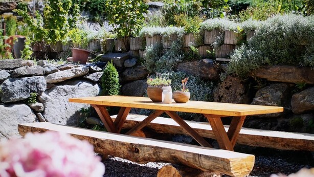 A wooden picnic table with benches, surrounded by a garden with various plants and a stone wall in the background. - Vacation apartment for up to 8 people (Latsch/Morter)