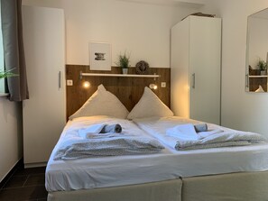 A neatly made bed with pillows and a blanket, a wooden headboard, a small shelf with decorative items, and a mirror on the wall. - Bernsteinsucher (Norden)