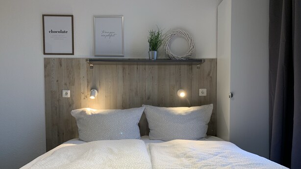 A neatly made bed with two pillows and two rolled towels, a wooden headboard, two wall-mounted lights, and two framed pictures on the wall. - Bernsteinsucher (Norden)
