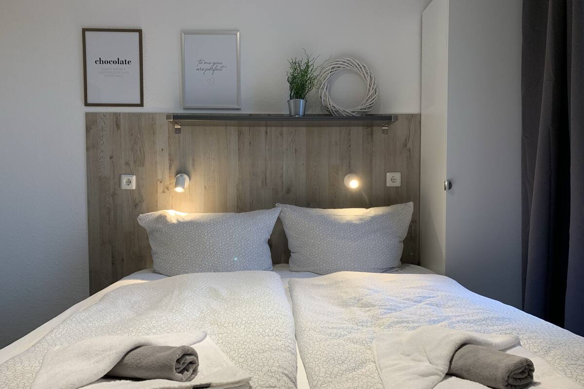 Una cama bien tendida con dos almohadas y dos toallas enrolladas, un cabecero de madera, dos lámparas fijadas a la pared y dos cuadros enmarcados.