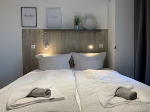A neatly made bed with two pillows and two rolled towels, a wooden headboard, two wall-mounted lights, and two framed pictures on the wall. - Bernsteinsucher (Norden)