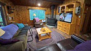 L’intérieur d’un chalet confortable, avec un canapé, un fauteuil, une table basse en bois, une cheminée, un téléviseur et une fenêtre donnant sur la forêt.