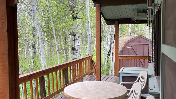 Une terrasse en bois avec une table ronde et des chaises, un tonneau en métal, et une vue sur des arbres et un bâtiment en arrière-plan.