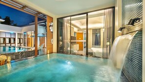 Un área de piscina interior moderna con una cascada, puertas de cristal que dan a una habitación con cama y baño, y un techo con tragaluces.
