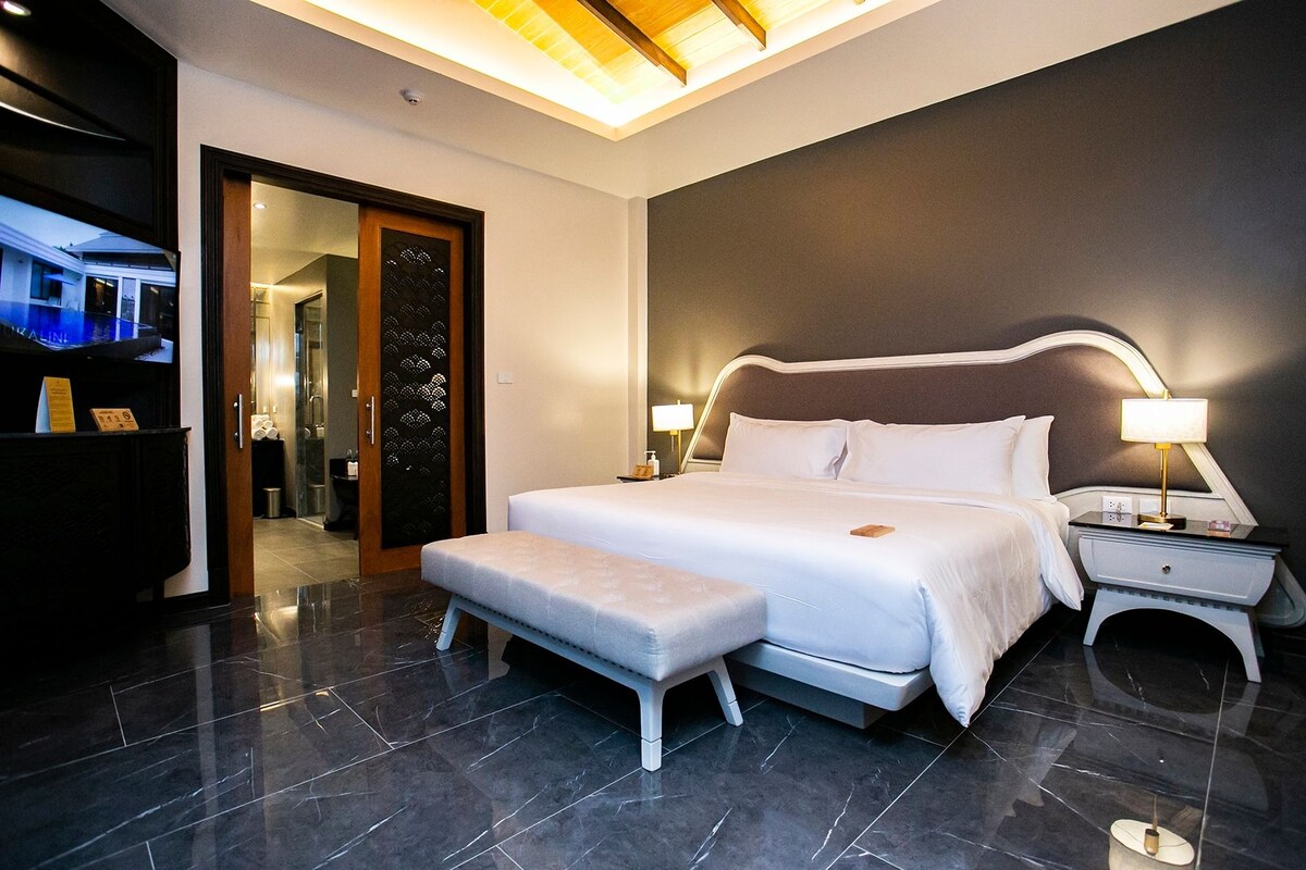 Quarto de hotel moderno com uma cama grande, mesas de cabeceira, um banco e uma porta para o banheiro.