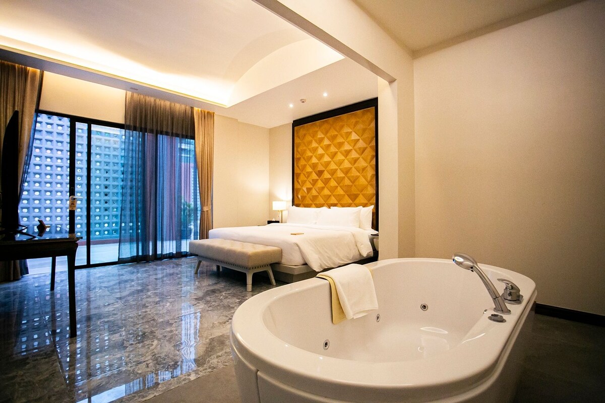 A modern hotel room with a large bathtub, a bed with a geometric-patterned headboard, and a view of a cityscape through the window.