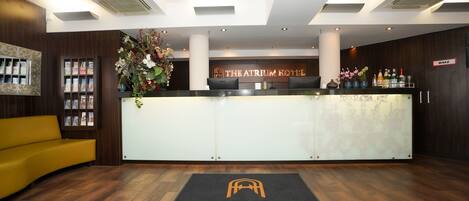 A reception area with a reception desk, a yellow sofa, a large floral arrangement, and a sign reading "THE ATRIUM HOTEL BY KML".