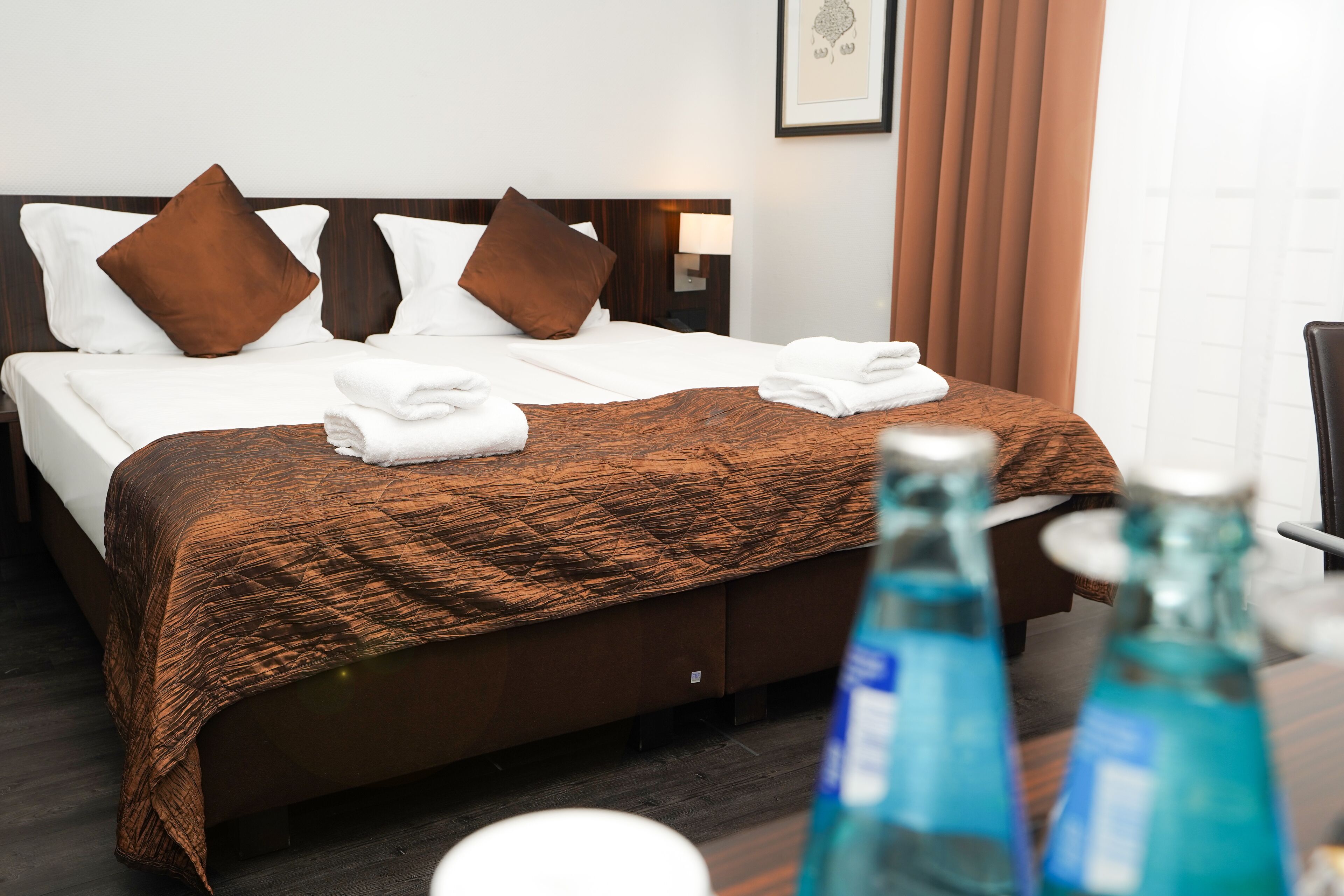 Habitación de hotel con una cama, ropa de cama marrón, almohadas blancas, cabecera marrón, mesita de noche con lámpara, ventana con cortinas y una mesa con dos botellas.