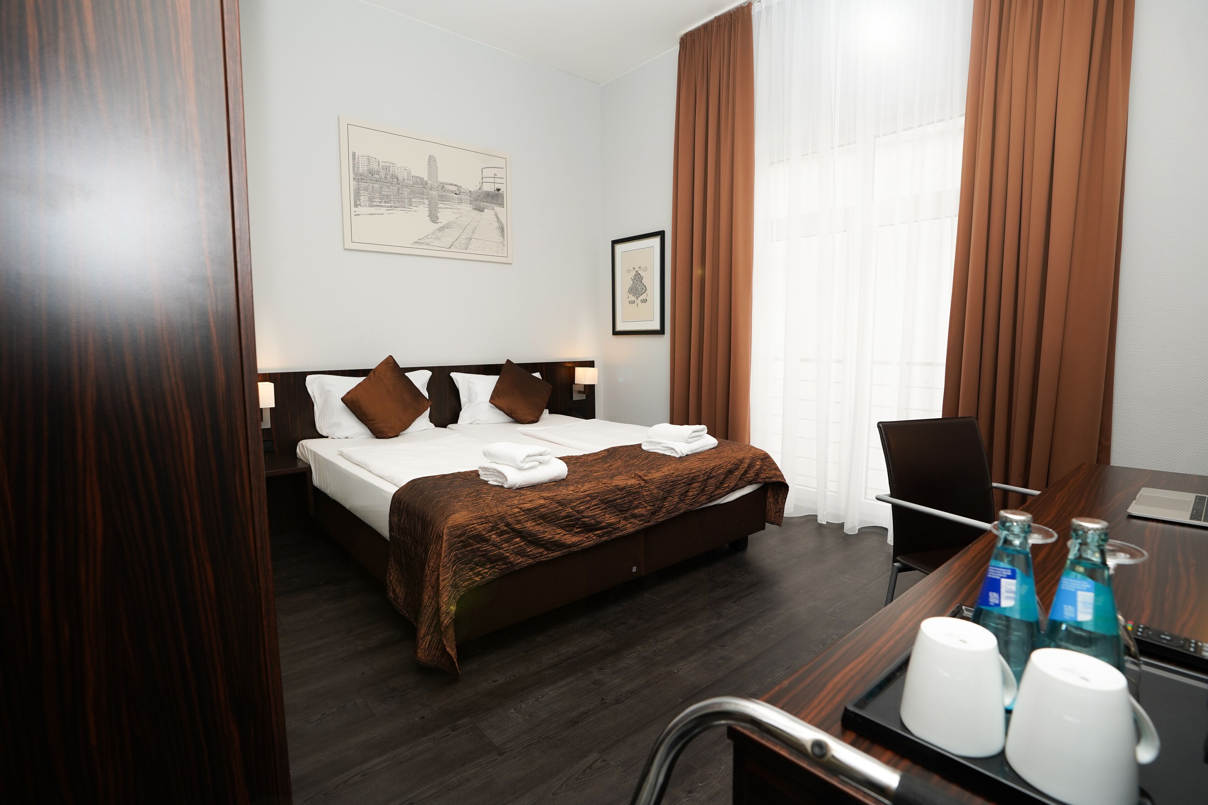 Una habitación de hotel con una cama, un escritorio con una computadora portátil y una silla. Hay dos botellas de agua y dos vasos sobre el escritorio.