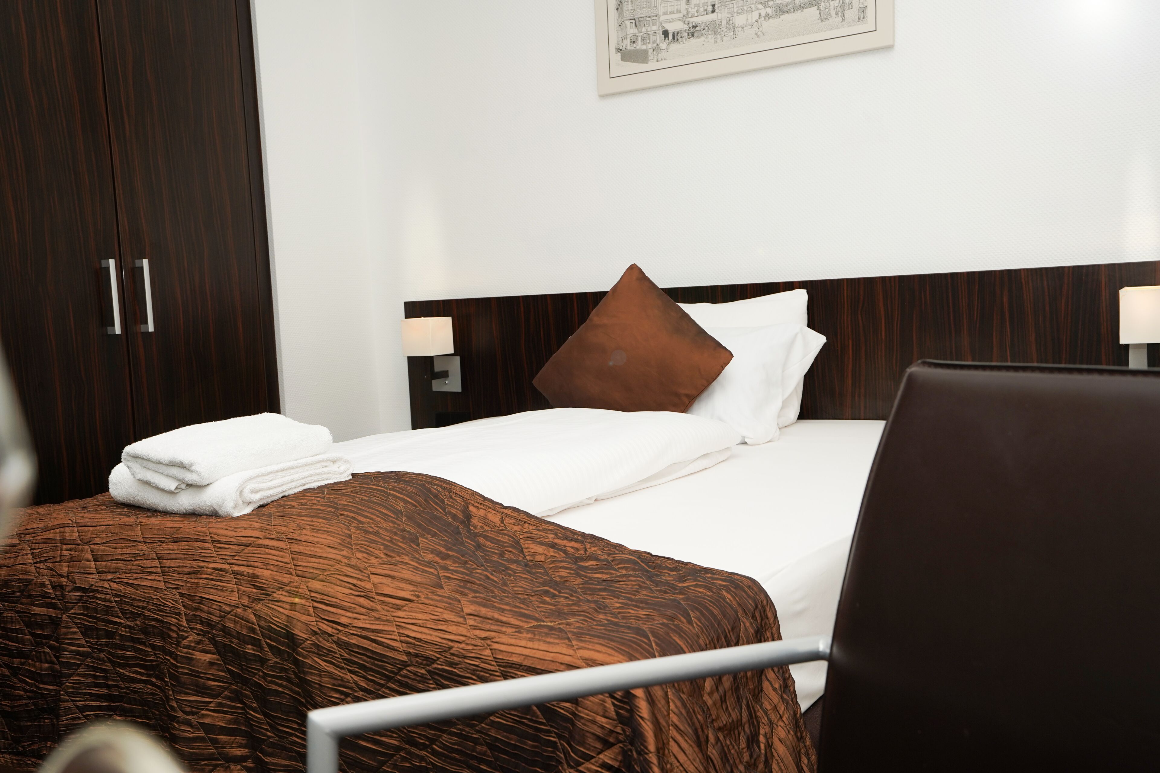 A hotel room with a neatly made bed, a brown quilt, a brown pillow, and a wooden wardrobe.
