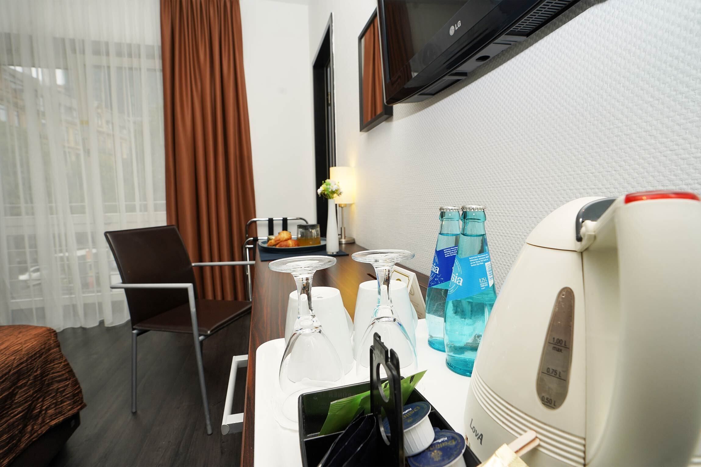A hotel room with a bed, a chair, a table with wine glasses, a water bottle, and a coffee maker.