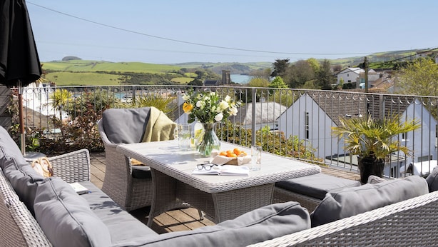 Ein Balkon mit Korbmöbeln, einem Tisch mit Blumen in einer Vase und Blick auf eine grüne Landschaft.