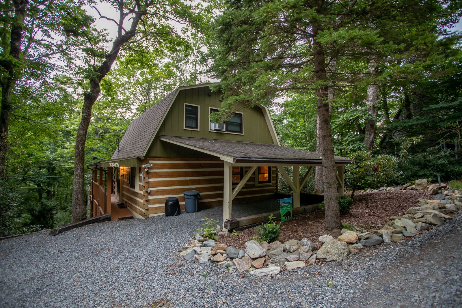 Une cabane rustique en rondins de bois, dotée d’un porche, entourée d’arbres et bordée d’une allée en gravier.
