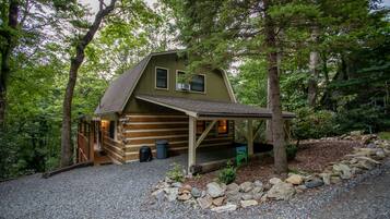 Une cabane rustique en rondins de bois, dotée d’un porche, entourée d’arbres et bordée d’une allée en gravier.