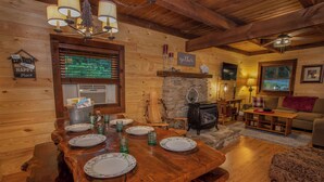 Un acogedor interior de cabaña con una mesa de comedor lista para cuatro comensales, una chimenea, paredes de madera y una zona de estar confortable.