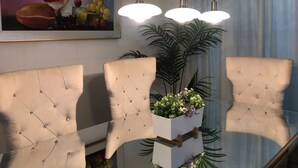 A dining area with a glass table, cushioned chairs, pendant lighting, a framed artwork, and a potted plant.