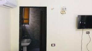 Ein Badezimmer mit Waschbecken, Spiegel, Wandklimaanlage, Lichtschalter und einem an der Wand befestigten Fernseher.