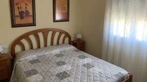 Une chambre à coucher avec un lit en bois, une table de chevet avec une lampe, deux tableaux encadrés au mur et une fenêtre avec des rideaux légers.