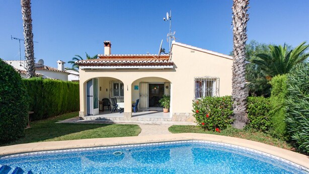 Vue d’une maison avec toit en tuiles, un patio couvert et une piscine, prise depuis le bord de la piscine.