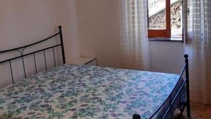 A single bed with a metal frame in a room with plain walls, a small window with curtains, and a ceiling light.