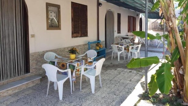 A covered outdoor seating area with white plastic chairs and a table, surrounded by a low wall and a large plant.