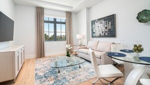 A modern living room with a glass coffee table, a sofa, a TV, and a dining area with a glass table and chairs. - SOCE FLATS 305 - Brand New Olde Naples Condo Steps to 5th Ave S! (Naples)