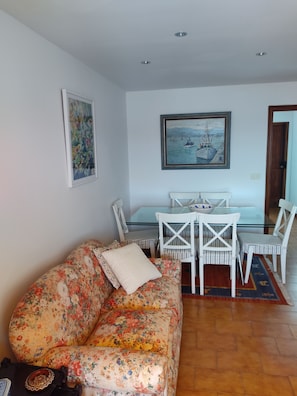 A living room with a floral sofa, a glass dining table with white chairs, a painting of boats, and a wooden door.