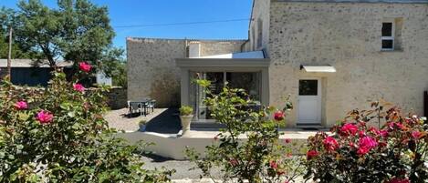 Un bâtiment en pierre de deux étages, doté d’un patio et d’un petit jardin agrémenté de fleurs roses.