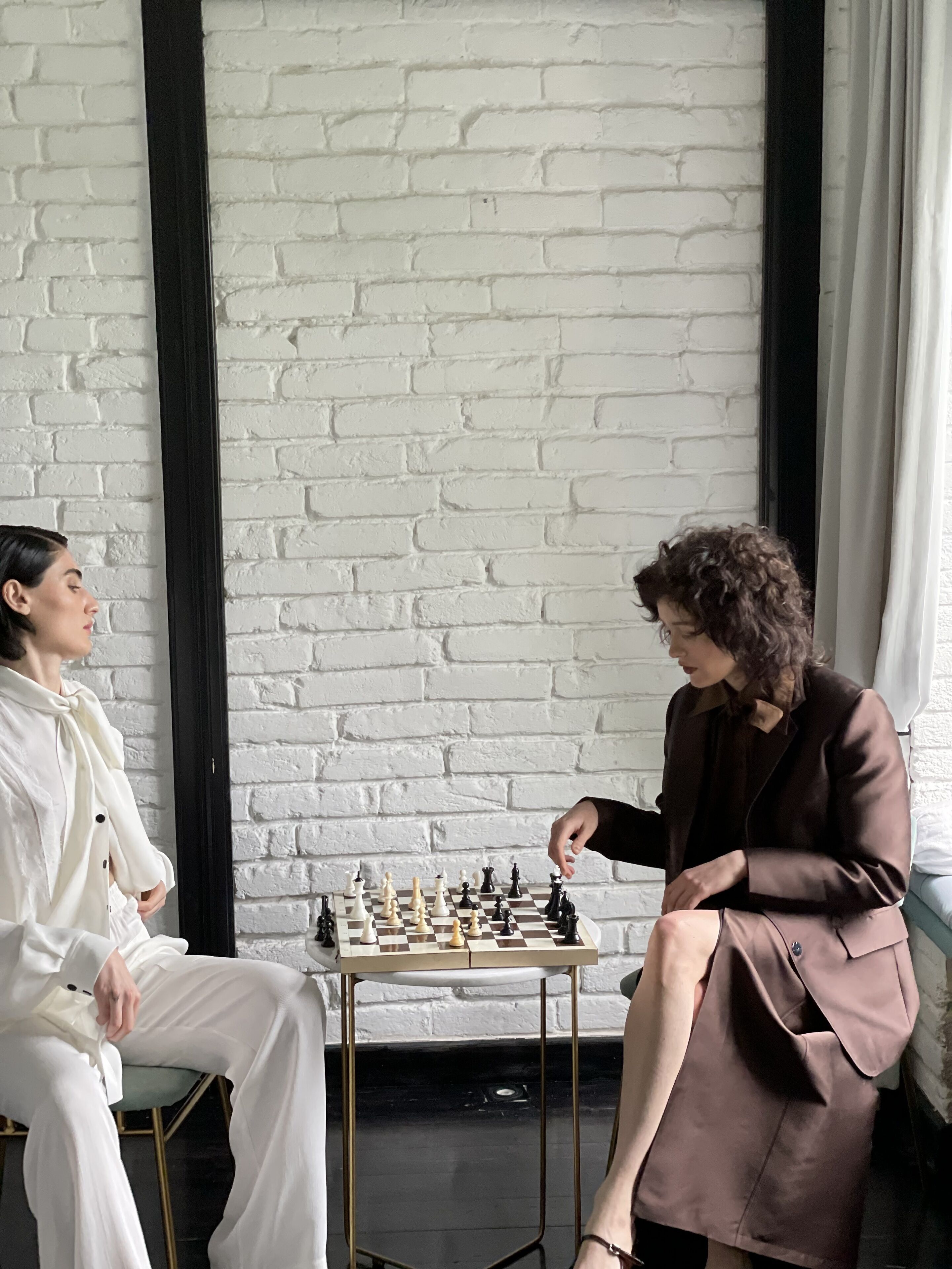 Two people playing chess in a room with a white brick wall and a window with curtains.
