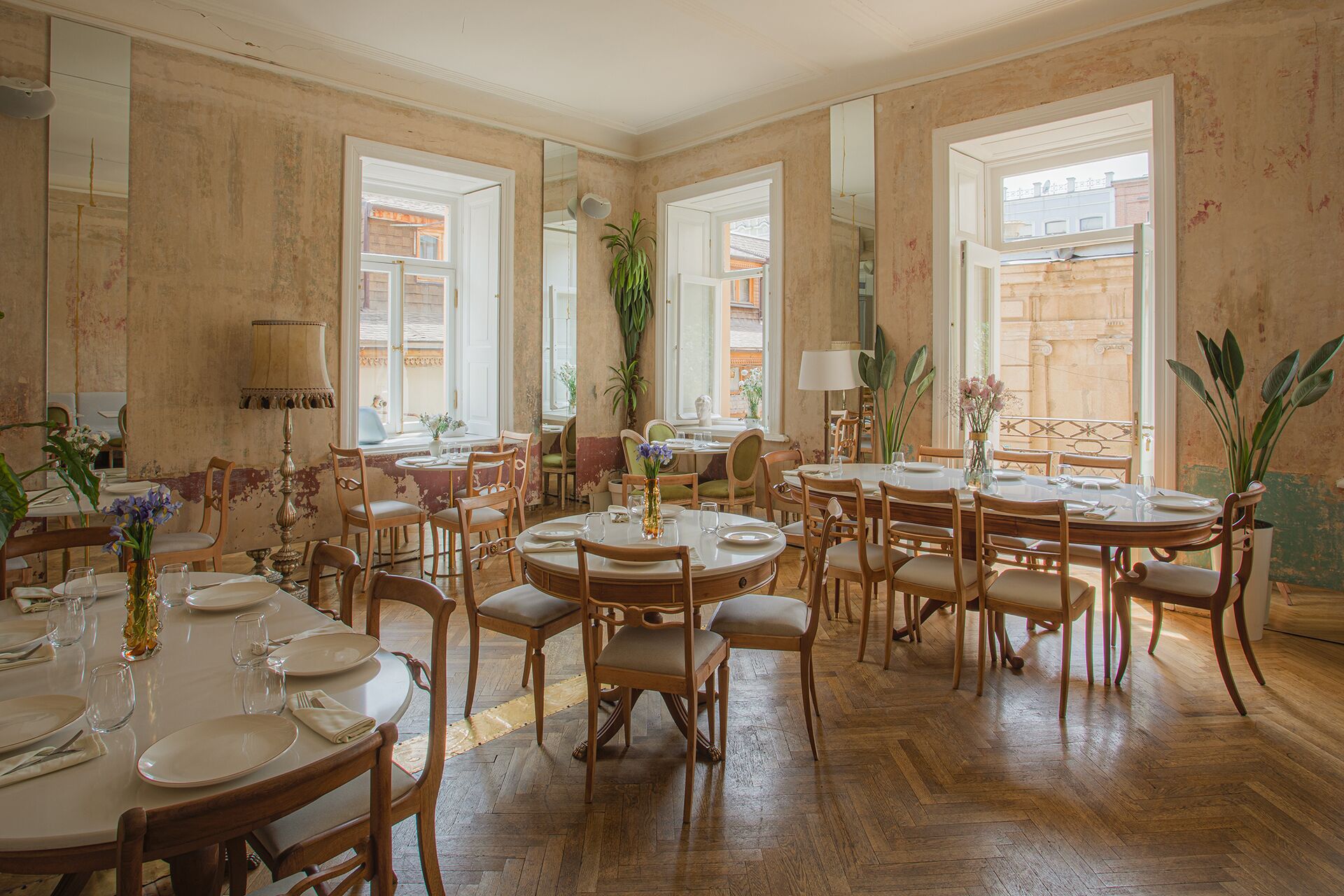 A dining area with multiple round tables and chairs, large windows, and potted plants.