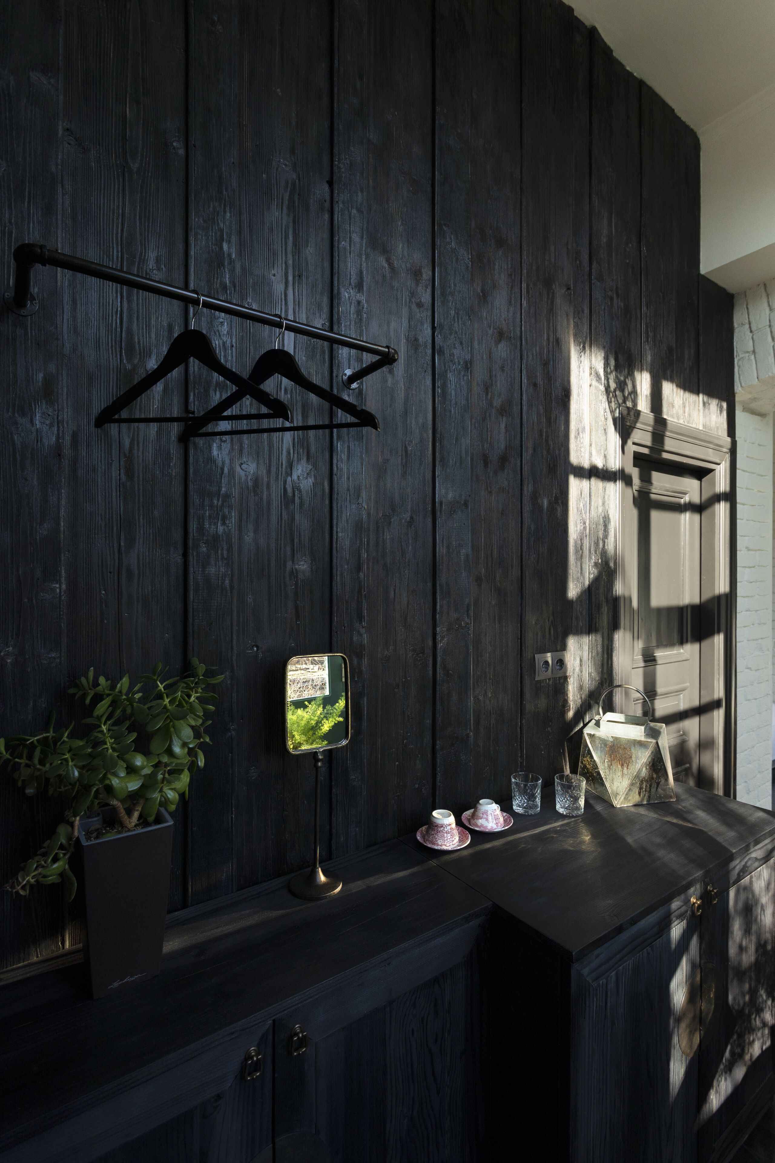 Una pared de madera oscura con un perchero metálico, un cuadro enmarco, una planta en maceta y una pequeña mesa con tazas y un vaso.