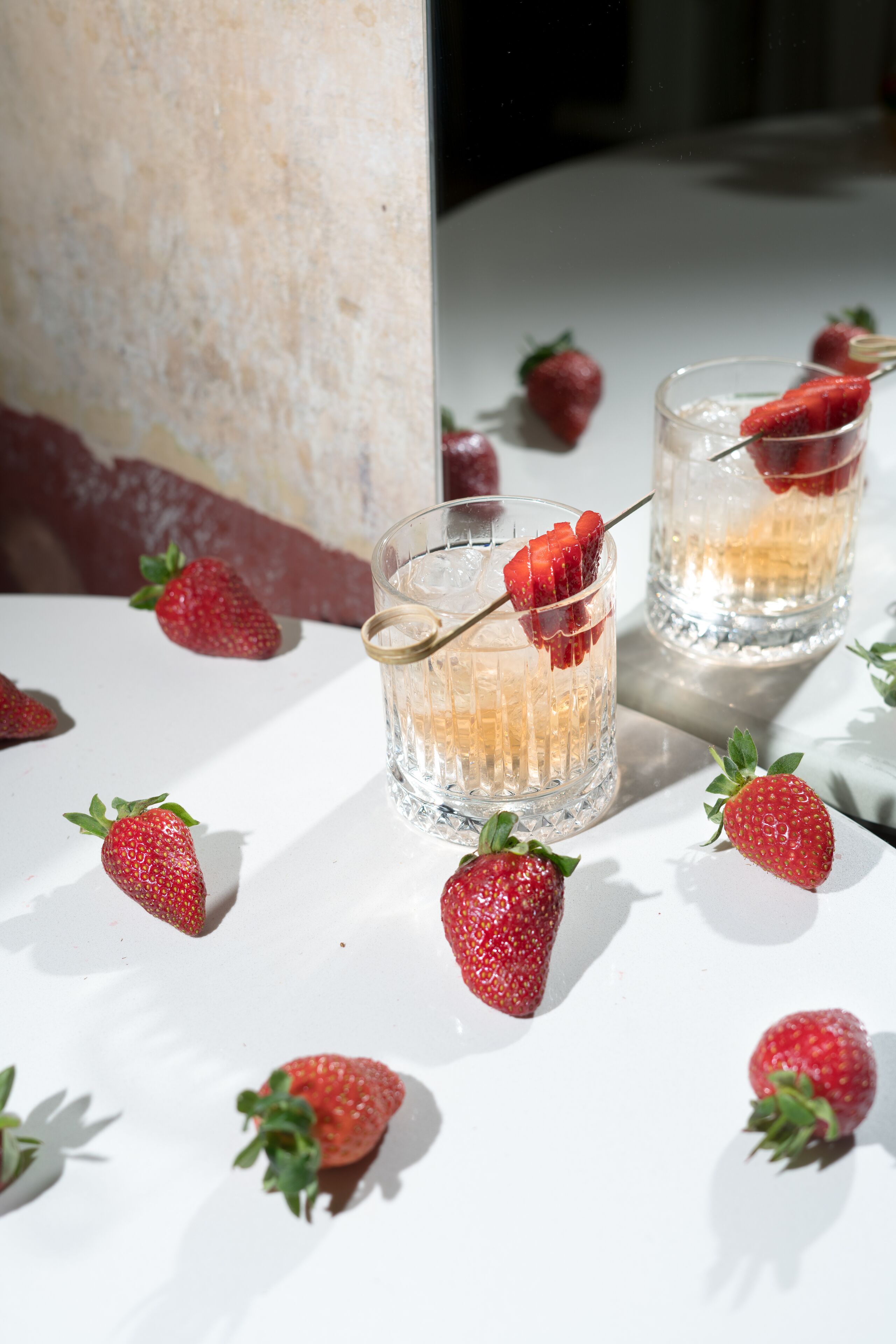 Two glasses of a beverage with strawberries, placed on a white surface with more strawberries scattered around.