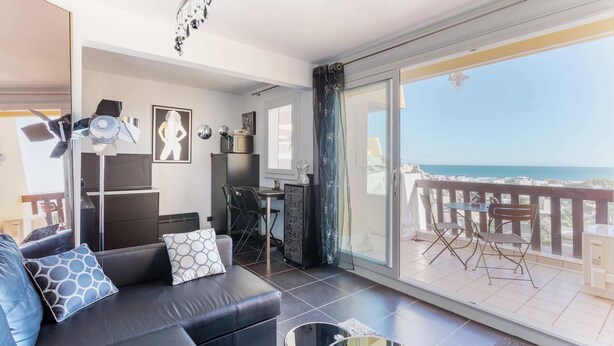A modern living room with a sofa, a dining table, and chairs. There is a balcony with a view of the sea.