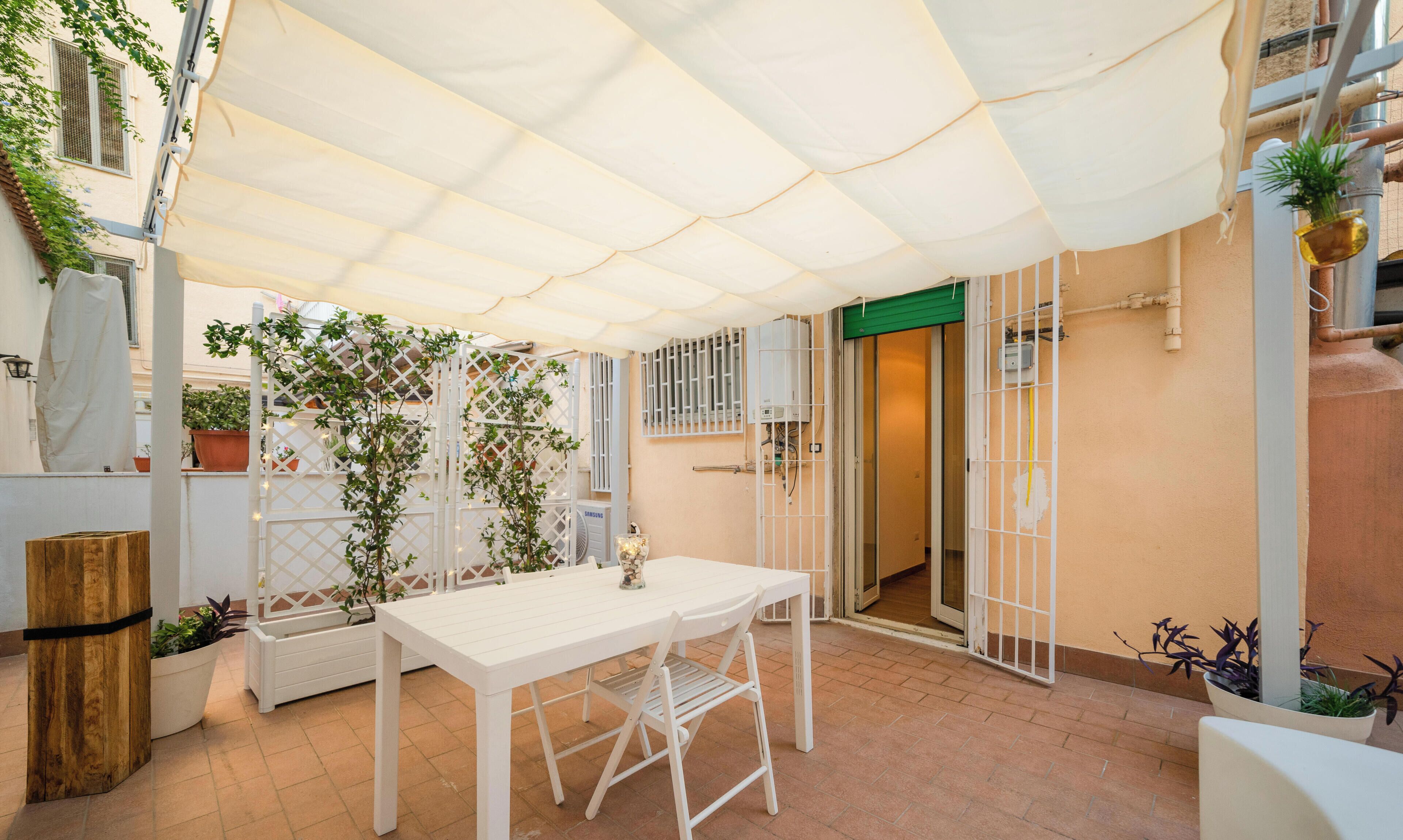 Un patio con una mesa y sillas blancas, rodeado de plantas en macetas y una cerca de celosía blanca.