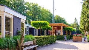 Un bâtiment avec un banc en bois portant l’inscription « BOSRAND », situé devant l’entrée, entouré de verdure et bordé d’un chemin pavé.