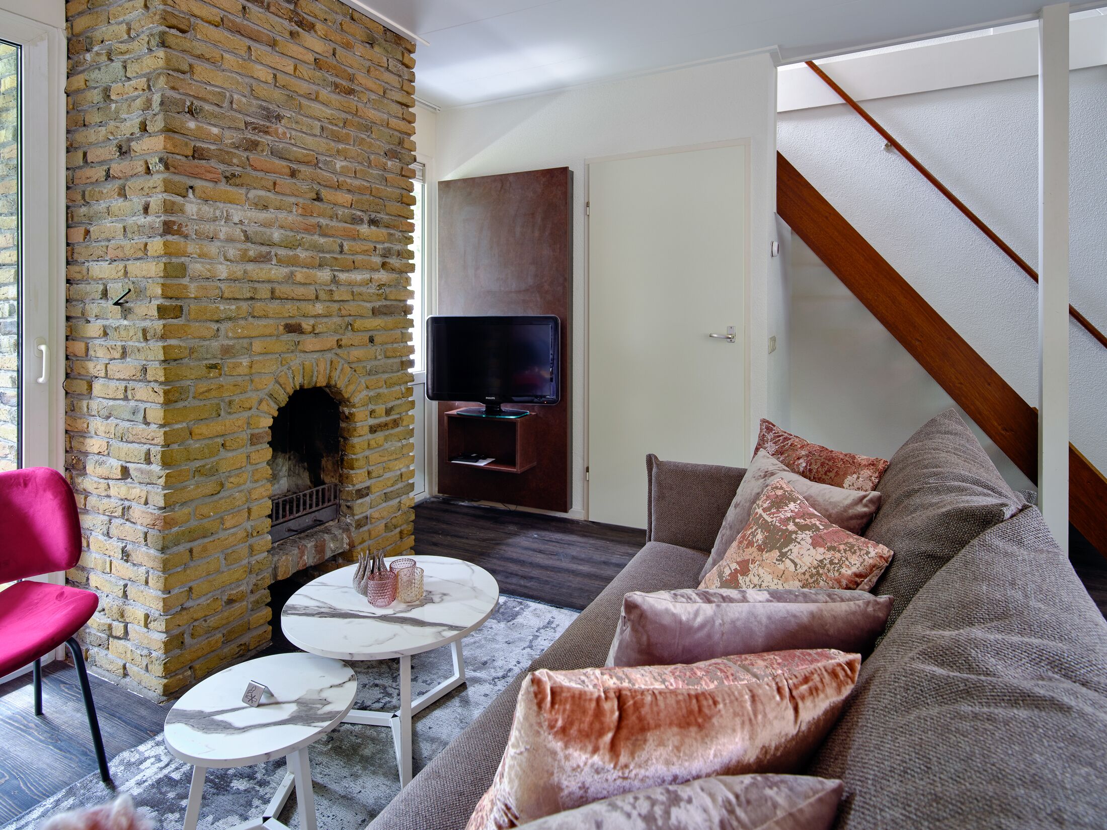 A living room with a brick fireplace, a sofa with patterned cushions, a television on a wooden stand, and a staircase.