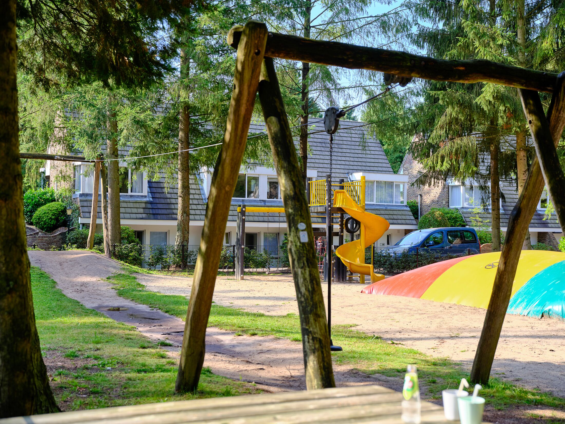 Ein Spielplatz mit einer gelben Rutsche und einer Schaukel, umgeben von Bäumen und einem Gebäude im Hintergrund.