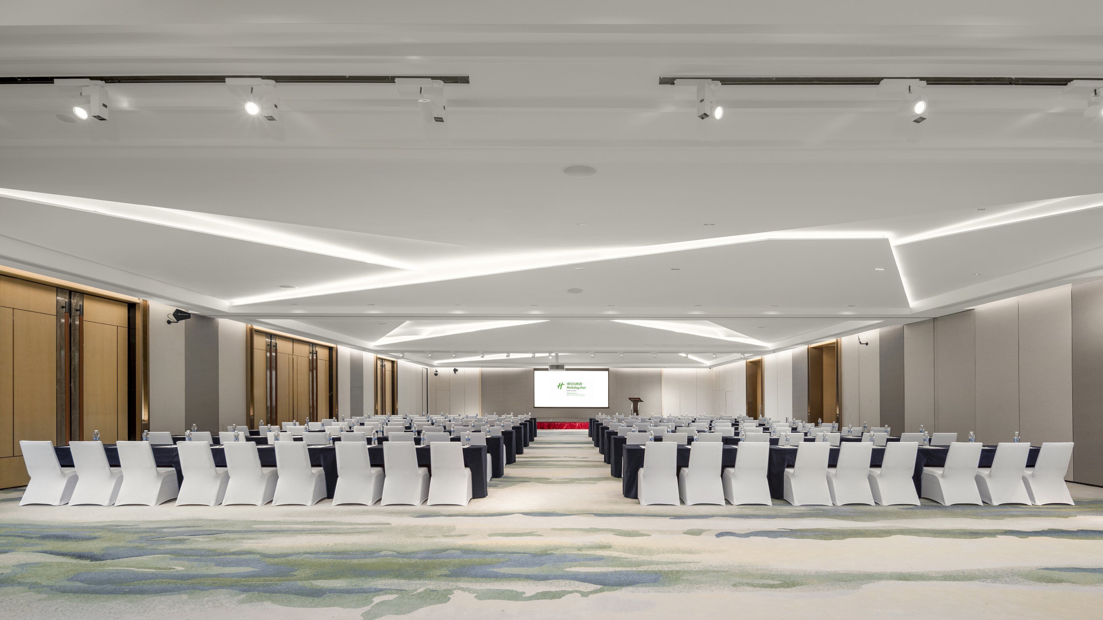 Una sala de conferencias con filas de sillas blancas, una pantalla grande al frente y piso alfombrado.