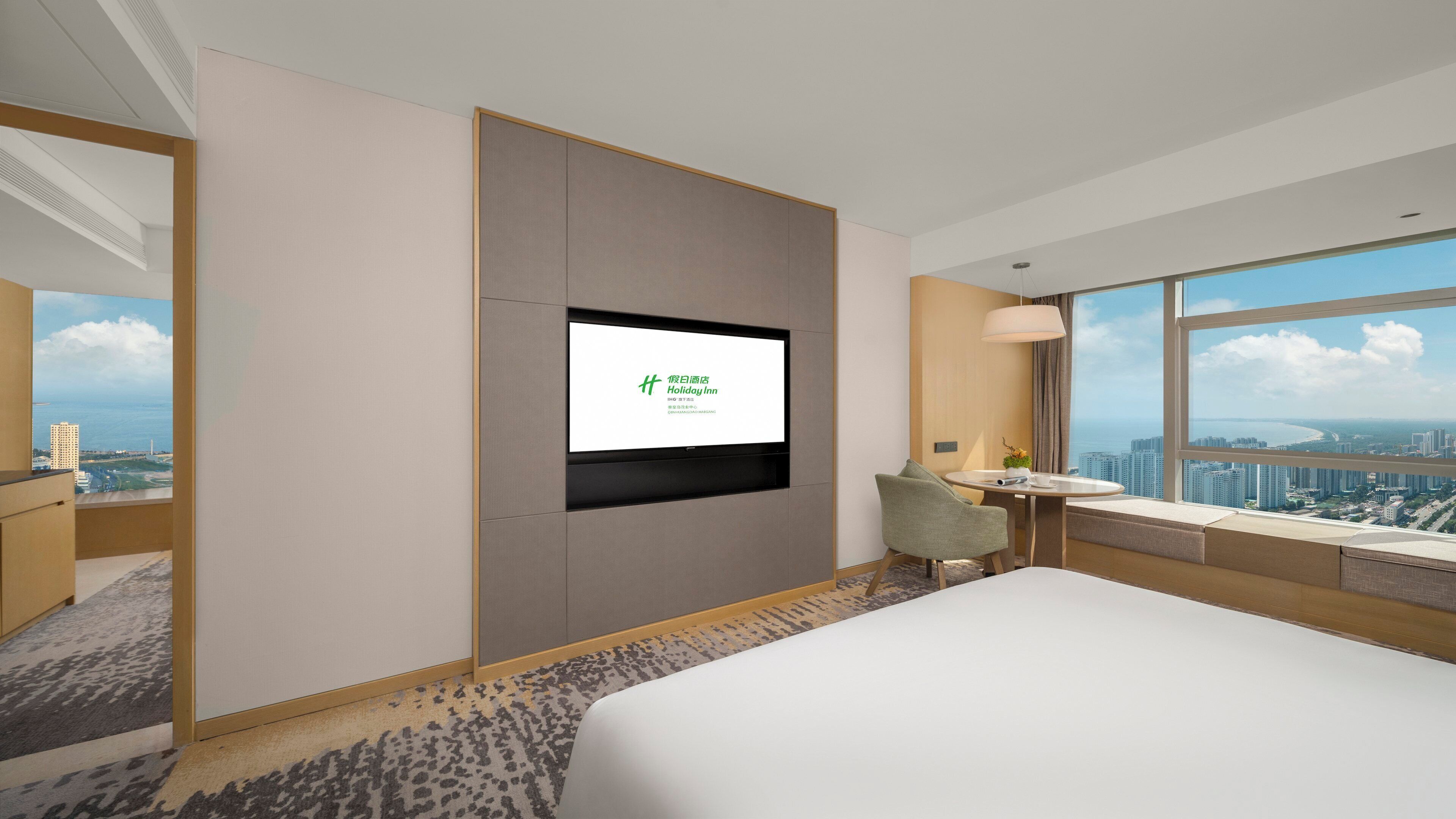 A hotel room with a bed, a desk with a chair, a television mounted on the wall, and a large window offering a city view.